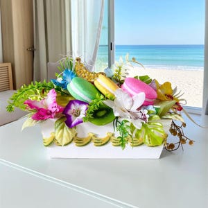 Può includere: Una fioriera rettangolare bianca decorata con un bordo di banane gialle, riempita con fogliame verde artificiale, vivaci orchidee rosa e bianche, una fetta di kiwi e tre colorati ornamenti a forma di macaron nei colori verde, giallo e rosa.