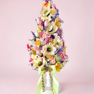 Può includere: Un albero di Pasqua decorativo composto da fiori colorati, uova e statuette di coniglietti. L'albero è sostenuto da una base a piedistallo bianca con un nastro verde chiaro e finiture dorate. L'albero è alto circa 46 cm.