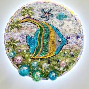 Può includere: Decorazione murale rotonda con un pesce colorato decorato con paillettes blu, gialle e verdi. L'opera include palme, stelle marine e bolle di vetro iridescenti, creando un tema sottomarino. Il diametro è di circa 30 cm.