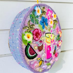Può includere: Un oggetto decorativo rotondo da appendere alla parete con un lato a righe blu e bianche e una parte anteriore testurizzata decorata con colorate applicazioni tropicali, tra cui una bevanda rosa, anguria, banana, cocco e vari fiori.