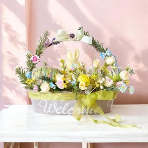 Può includere: Un cestino pasquale con un design grigio e verde, pieno di fiori colorati, un coniglietto giallo e uova decorative. Il cestino ha un manico ornato da un fiocco bianco e la parola "Welcome" in corsivo.