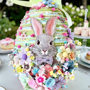 Könnte beinhalten: Osterei-förmige Dekoration mit einem grauen Hasengesicht und einer lila Fliege. Das Ei ist mit bunten Blumen, Bändern und einer grünen Schleife verziert. Der Hintergrund ist verschwommen, was auf eine Umgebung im Freien hindeutet.