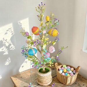 Könnte beinhalten: Ein Osternbaum mit bunten, garnumwickelten Eiern und zarten Blüten. Der Baum steht in einem gewebten Topf auf einem Holztisch. Ein kleiner Korb mit pastellfarbenen Eiern steht neben dem Baum.