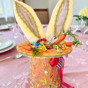 Könnte beinhalten: Ein skurriler Osterhut mit großen, flauschigen gelben Hasenohren. Der pfirsichfarbene Hut ist mit floralen Stickereien, Miniaturkarotten, Blumen und einem dekorativen Band verziert. Der Hut steht auf einer rosa Tischdecke.