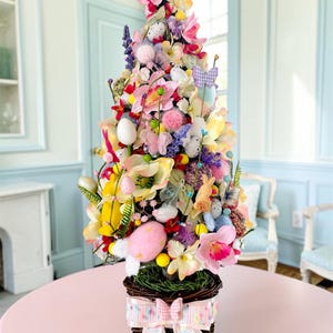 Può includere: Una composizione decorativa a forma di albero pasquale in un cestino di vimini, traboccante di fiori artificiali colorati, uova, farfalle e piccole figure di coniglietti. La composizione è posta su un tavolo rosa, con un nastro decorativo legato attorno al cestino.
