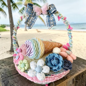Puede incluir: Una cesta decorativa con un cono de helado de tela, un lazo de tweed azul con un detalle de mariposa rosa y varias flores de tela en tonos azules, verdes y rosas. El asa está envuelta en hilo de colores y tiras de tela deshilachadas.