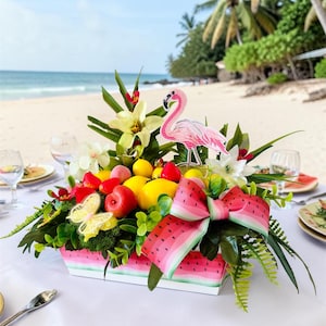 Peut inclure: Un centre de table rectangulaire avec une base et un ruban à motif pastèque, rempli de citrons artificiels, de pommes rouges, de feuillage vert, de lys blancs, d'une découpe de flamant rose et d'un papillon jaune, sur une nappe blanche.