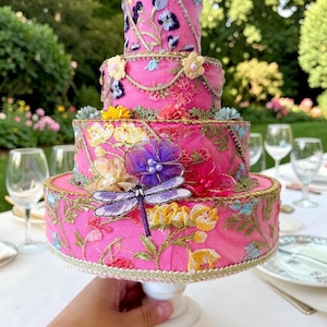 Può includere: Una torta decorativa rosa a tre livelli con ricami floreali e un accento di libellula. La torta è adornata con fiori colorati e finiture dorate e poggia su un piedistallo bianco. La torta è alta circa 20 cm.
