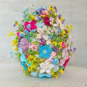 Könnte beinhalten: Eine farbenfrohe Ostereier-Dekoration aus Stroh, Bändern und künstlichen Blumen. Das Ei ist mit rosa, blauen, gelben und weißen Blumen sowie Bändern und Schleifen verziert.