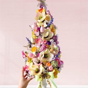 Puede incluir: Un árbol de Pascua decorativo adornado con flores coloridas, huevos y figuras de conejitos. El árbol está sobre un pedestal blanco y adornado con una cinta verde claro. El árbol mide aproximadamente 46 cm de alto.