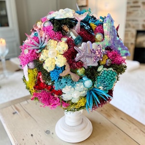 Può includere: Una composizione decorativa sferica su una base bianca, caratterizzata da muschio vivace nei toni del rosa, giallo e verde, arricchita da fiori di paillettes, stelle marine, code di sirena e una piccola bottiglia di vetro con un rotolo.