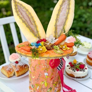 Può includere: Un cappello pasquale stravagante con grandi orecchie da coniglio di peluche. Il cappello color pesca è adornato da ricami floreali, carote in miniatura e fiori colorati. Un nastro rosso con un accento a scacchi aggiunge un tocco giocoso. Torte e pasticcini sullo sfondo.