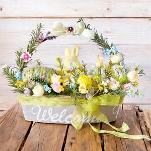 Könnte beinhalten: Ein dekorativer Osterkorb gefüllt mit bunten Blumen, einem gelben Hasen und einem Wollknäuel. Der graue Korb hat eine grüne Umrandung und den Schriftzug "Welcome" in weißer Schrift. Ein grünes Band ist um den Korb gebunden.