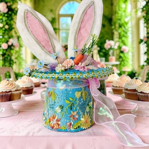 Può includere: Un cappello pasquale stravagante con grandi orecchie da coniglio bianche con interni rosa e finiture argentate. Il cappello blu è adornato da ricami floreali colorati e una carota. Un nastro rosa trasparente aggiunge un tocco delicato. Cupcake sullo sfondo.