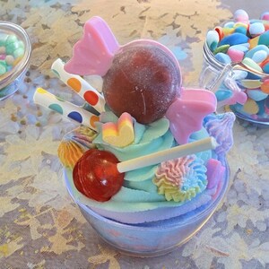 May include: A colorful dessert creation in a clear bowl, featuring pastel-colored frosting, a red lollipop, and various candies. The arrangement includes a large brown candy, pink bow-shaped candies, and decorative straws with polka dots. Other candies are visible in the background.