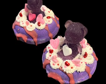 Könnte beinhalten: Zwei violette Badebomben mit einem Teddybären oben drauf. Die Badebomben haben weiße Glasur, rosa und rote herzförmige Dekorationen und rosa Tröpfchen. Der Hintergrund ist schwarz.