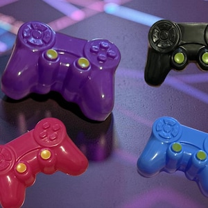 May include: Four colorful game controllers in purple, black, red, and blue. Each controller has a glossy finish with yellow buttons and a circular directional pad. The controllers are arranged on a purple surface.