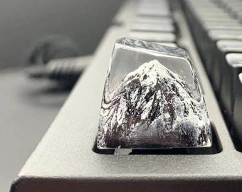 May include: A custom keyboard keycap featuring a miniature mountain scene encased in clear resin. The mountain is detailed with white snow and dark rock, set against a black keyboard.