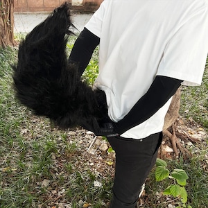 May include: A black furry tail costume accessory. The tail is long and bushy, with a realistic look. It is worn by a person wearing a white t-shirt and black pants.