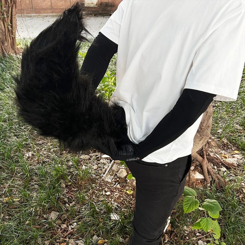 May include: A black furry tail costume accessory. The tail is long and bushy, with a realistic look. It is worn by a person wearing a white t-shirt and black pants.