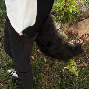 May include: A person wearing black pants and a white shirt with a black furry tail attached to the back of their waist.