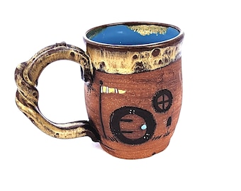 May include: A brown ceramic mug with a unique, twisted handle. The mug features a hand-painted design of a small house with a blue door and a yellow flag.