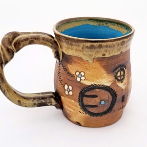 Puede incluir: Una taza de cerámica marrón con un asa retorcida. La taza está decorada con una escena pintada de una pequeña casa con una puerta redonda y una ventana. La casa está rodeada de flores blancas.