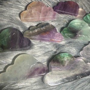 May include: A collection of cloud-shaped fluorite stones in various colors, including purple, green, and clear. The stones are polished and have a smooth surface. The clouds are arranged on a gray, fuzzy surface.