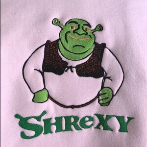 May include: A pink sweatshirt featuring an embroidered image of Shrek in green, wearing a brown vest, with the word "Shrexy" in green underneath.