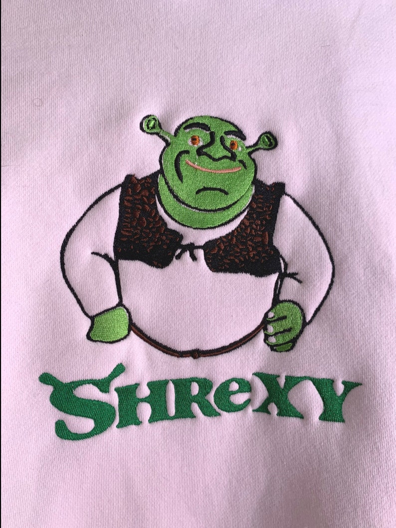 May include: A pink sweatshirt featuring an embroidered image of Shrek in green, wearing a brown vest, with the word "Shrexy" in green underneath.