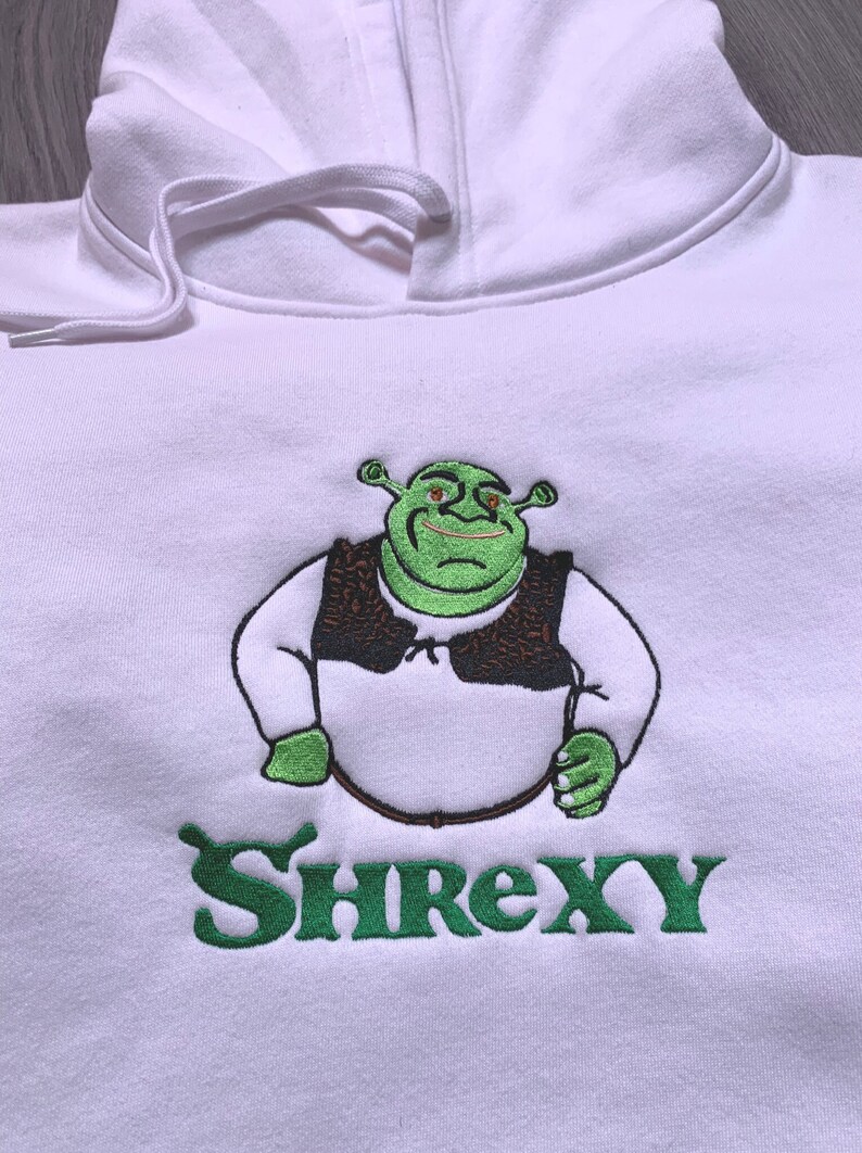 May include: White hoodie featuring an embroidered green image of Shrek wearing a brown vest and the text "Shrexy" in green underneath.