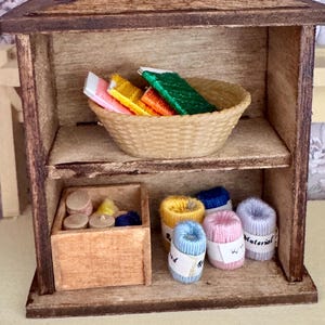 Pode incluir: Uma estante de madeira em miniatura com dois níveis. A prateleira superior contém uma pequena cesta trançada cheia de carretéis de linha coloridos. A prateleira inferior contém uma caixa de madeira com linha e vários carretéis individuais em azul, rosa e amarelo.