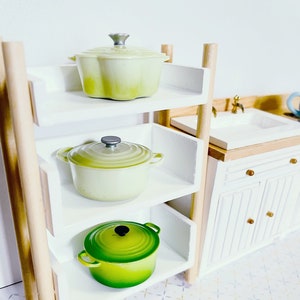 May include: A white wooden shelf unit with three shelves, each holding a different coloured cast iron pot. The top shelf has a light green pot, the middle shelf has a light green pot, and the bottom shelf has a green pot.