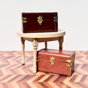 May include: Two miniature leather suitcases with gold accents and clasps, one brown and one red, on a wooden table with turned legs. The table is on a wood floor with a herringbone pattern.