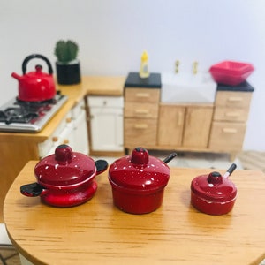 May include: Three miniature red pots with white speckles and black handles. The pots are arranged in a row on a wooden surface.