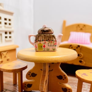 Può includere: Una teiera in miniatura rosa e marrone a forma di casa con un tetto di paglia e una porta verde. La teiera è appoggiata su un tavolo di legno con gambe a forma di zampa d'orso.