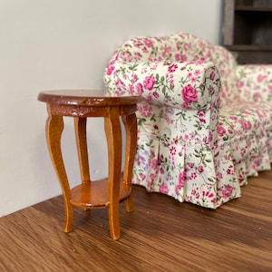 May include: A small, wooden end table with a round top and three legs. The table is painted a light brown color and has a floral pattern on the top. The table is sitting in front of a floral patterned sofa.