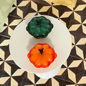 Puede incluir: Dos pequeños platos de cerámica con forma de calabaza, uno verde y otro naranja, sobre un pedestal blanco.