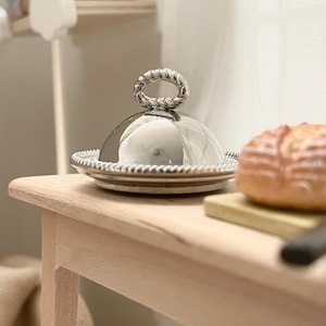 Puede incluir: Un plato de servir plateado con una tapa abovedada y un asa con forma de cuerda. El plato está sobre una mesa de madera con una hogaza de pan y un cuchillo.