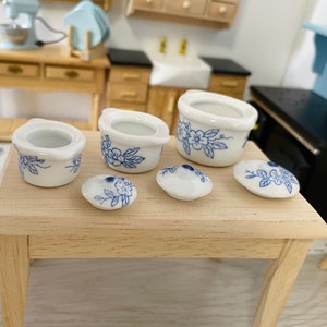 May include: A set of five miniature white ceramic pots with blue floral designs. The pots are arranged on a light wood table.