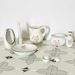 May include: A set of miniature white porcelain bathroom accessories. The collection features a jug, bowl, soap dish, and other items, all decorated with pink rose floral designs and gold trim. The items are arranged on a patterned tile surface.