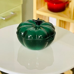 May include: A green pumpkin-shaped cast iron Dutch oven with a black knob on the lid. The oven is sitting on a white table.