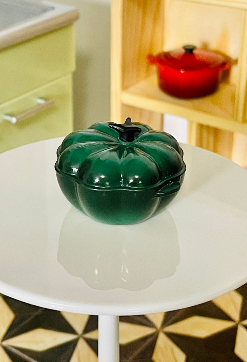 May include: A green pumpkin-shaped cast iron Dutch oven with a black knob on the lid. The oven is sitting on a white table.