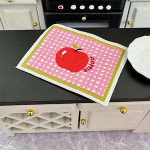 Może przedstawiać: Miniaturowa mata kuchenna z różowym wzorem w kratkę i czerwonym wzorem jabłka. Mata ma zieloną obwódkę, a z boku napis "apple". Ta dekoracyjna mata to uroczy dodatek do domku dla lalek lub miniaturowej kuchni.