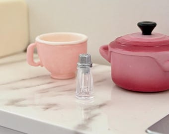 May include: A miniature kitchen scene with a pink teacup, a small pink pot with a black handle, and a clear salt shaker with a silver top. The items are on a white countertop with a marble pattern.