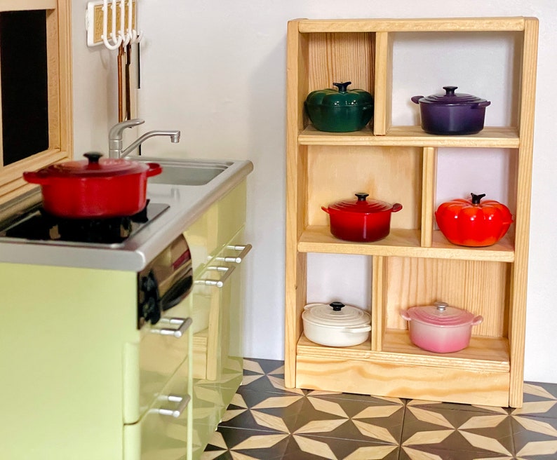 May include: A wooden shelf unit with six shelves, each holding a different colored cast iron pot. The pots are red, green, white, pink, orange, and purple.