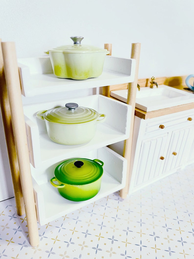 May include: A white wooden shelf unit with three shelves, each holding a different coloured cast iron pot. The top shelf has a light green pot, the middle shelf has a light green pot, and the bottom shelf has a green pot.