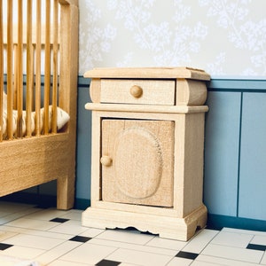 May include: A small wooden nightstand with a single drawer and a cabinet door. The nightstand is painted in a light wood color and has a simple design. It is sitting on a tiled floor.