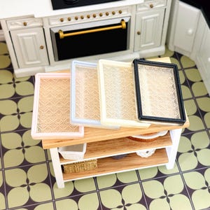 May include: Miniature dollhouse kitchen scene featuring a white oven and a wooden table. The table holds four decorative trays with patterned glass inserts and colored frames: pink, light blue, cream, and black. A small white basket and a woven basket are on the lower shelf.