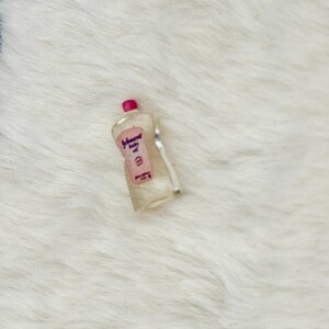 Może przedstawiać: Miniaturowa butelka z przezroczystego plastiku z oliwką dla dzieci Johnson's, posiadająca r&oacute;żową etykietę z napisem Johnson's baby oil wykonanym ciemną czcionką oraz małą r&oacute;żową nakrętkę. Butelka ma zakrzywiony kształt i jest wykonana jako replika.